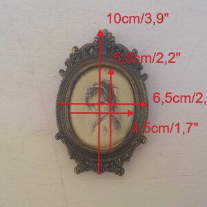 May include: A small oval-shaped brass picture frame with an ornate design. The frame is 10 cm tall and 6.5 cm wide. The frame holds a black and white drawing of a woman's face.
