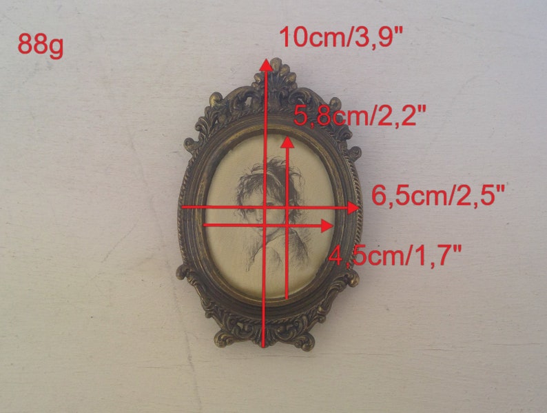 May include: A small oval-shaped brass picture frame with an ornate design. The frame is 10 cm tall and 6.5 cm wide. The frame holds a black and white drawing of a woman's face.
