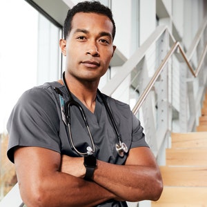May include: A man wearing a gray medical scrub top with a stethoscope around his neck. He is standing in front of a staircase with his arms crossed and a watch on his wrist.