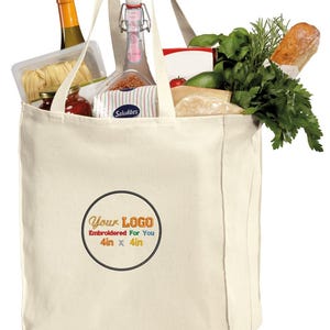 May include: A natural canvas tote bag with a large, round, black circle containing the text "Your LOGO Embroidered For You 4in x 4in". The bag is filled with groceries, including a bottle of wine, a loaf of bread, and fresh herbs.