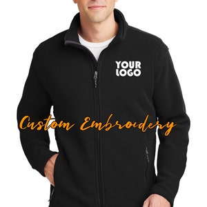 May include: A black fleece jacket with a zipper closure and two front pockets. The jacket has a white embroidered logo that says "YOUR LOGO" on the left chest.