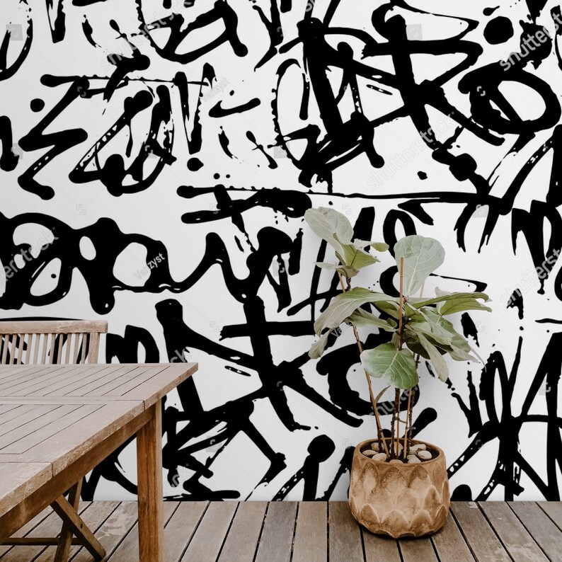 May include: Black and white abstract art print with a black and white plant in a brown pot in front of it. The print is on a white wall and there is a wooden table and chair in front of it.