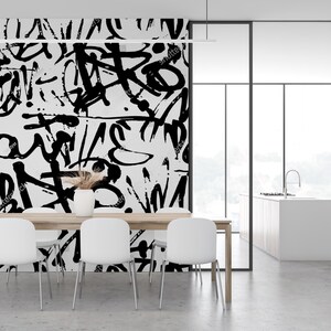 May include: A dining room with a white table and chairs. The wall behind the table is covered in a black and white abstract graffiti pattern. The room has a large window and a view of a kitchen.