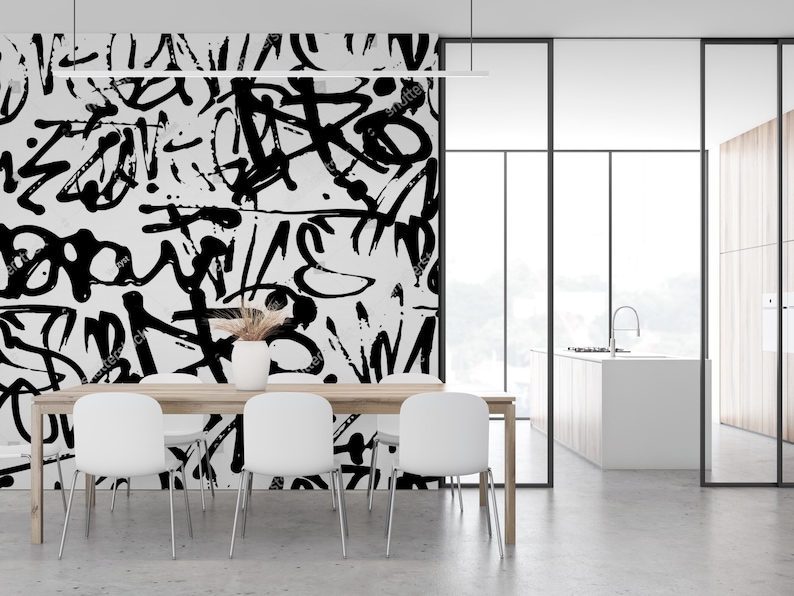 May include: A dining room with a white table and chairs. The wall behind the table is covered in a black and white abstract graffiti pattern. The room has a large window and a view of a kitchen.