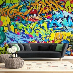 Puede incluir: Un vibrante mural de graffiti en tonos amarillos, azules, verdes y naranjas domina la pared detr&aacute;s de un sof&aacute; negro. La habitaci&oacute;n est&aacute; decorada con una alfombra neutra, dos pufs y una planta en maceta, creando un espacio de vida moderno.