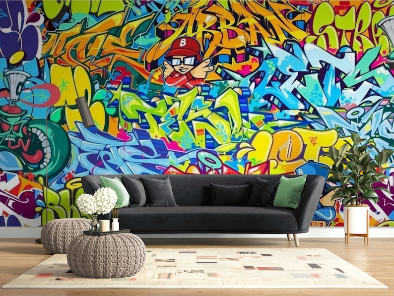Puede incluir: Un vibrante mural de graffiti en tonos amarillos, azules, verdes y naranjas domina la pared detr&aacute;s de un sof&aacute; negro. La habitaci&oacute;n est&aacute; decorada con una alfombra neutra, dos pufs y una planta en maceta, creando un espacio de vida moderno.