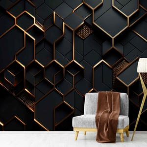 May include: A modern geometric wall mural in black and gold. The design features a 3D effect with geometric shapes and gold detailing. A grey chair with a brown blanket and a gold floor lamp are in front of the wall.