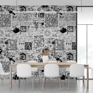 May include: A dining room with a light wood table and white chairs. The wall is covered in black and white wallpaper with script and decorative letter designs. A white vase with dried plants sits on the table.