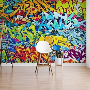 Puede incluir: Un vibrante mural de graffiti con letras abstractas coloridas y personajes de dibujos animados. La obra de arte incluye tonos de azul, amarillo, rojo y verde. Una escalera de madera, una silla blanca y una planta en maceta est&aacute;n frente al mural.