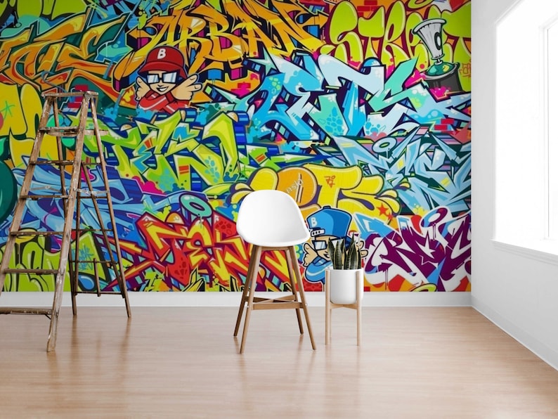 Puede incluir: Un vibrante mural de graffiti con letras abstractas coloridas y personajes de dibujos animados. La obra de arte incluye tonos de azul, amarillo, rojo y verde. Una escalera de madera, una silla blanca y una planta en maceta est&aacute;n frente al mural.