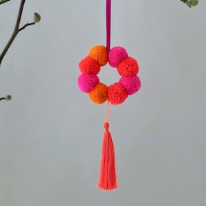 Könnte beinhalten: Ein rosafarbener und orangefarbener Pompon-Kranz mit einem rosafarbenen Band und einem leuchtend orangen Quasten, der an einem Ast hängt.