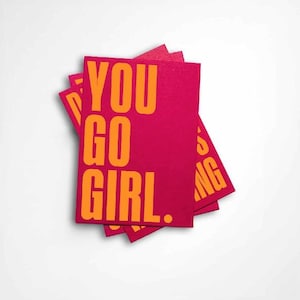 Può includere: Una pila di biglietti rosa con le parole "YOU GO GIRL" in grassetto arancione. I biglietti sono leggermente a ventaglio, su sfondo bianco.