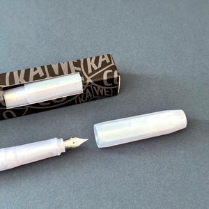 以下が含まれることがあります： 銀色のペン先とキャップが付いた、白く光る万年筆。黒と白の箱が横に置かれています。箱にはブランド名「KAWECO」が印刷されています。ペンは開いています。