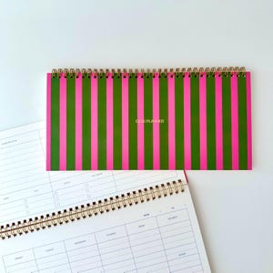 Könnte beinhalten: Ein horizontaler Schreibtischplaner mit goldener Spiralbindung und einem lebendigen Muster aus abwechselnden vertikalen pinken und waldgrünen Streifen. Die Worte DESK PLANNER sind in kleinen goldenen Großbuchstaben in der Mitte aufgedruckt.