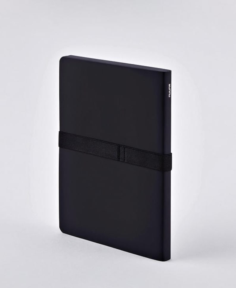 May include: A black notebook with a black elastic band closure. The notebook has a minimalist design and a smooth, matte finish. The word "nuuna" is printed vertically on the spine.