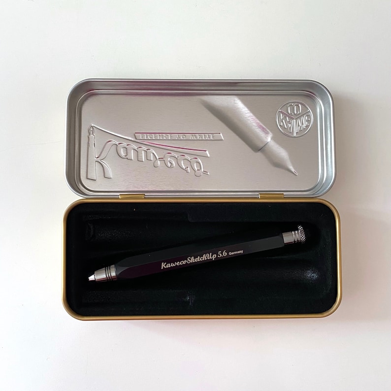May include: A silver metal tin with a black interior. The tin is open and contains a black mechanical pencil with silver accents. The pencil has the text "Kaweco SketchUp 5.6 Germany" printed on it.