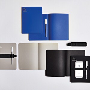 May include: An assortment of notebooks in blue, gray, and black, some open to reveal blank pages. Each notebook has a pen and a black elastic band. The text "NOT WHITE" is printed on some of the notebooks.