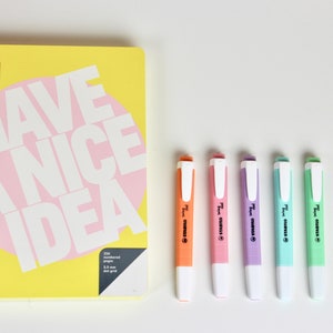 以下が含まれることがあります： 「HAVE A NICE IDEA」の白文字とピンクの円が描かれた黄色のノートと、5色の蛍光ペン。蛍光ペンはオレンジ、ピンク、紫、水色、緑で、白いキャップが付いています。