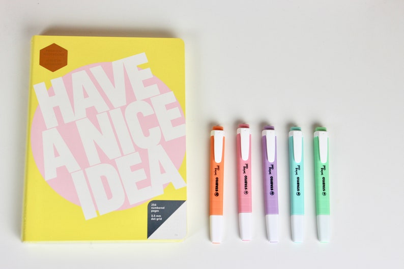 以下が含まれることがあります： 「HAVE A NICE IDEA」の白文字とピンクの円が描かれた黄色のノートと、5色の蛍光ペン。蛍光ペンはオレンジ、ピンク、紫、水色、緑で、白いキャップが付いています。