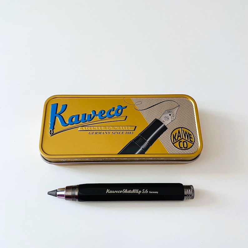 May include: A black mechanical pencil with the text "Kaweco SketchUp 5.6 Germany" on the side. The pencil is in a gold tin with the text "Kaweco License to Write Germany Since 1883" and a black and silver fountain pen.