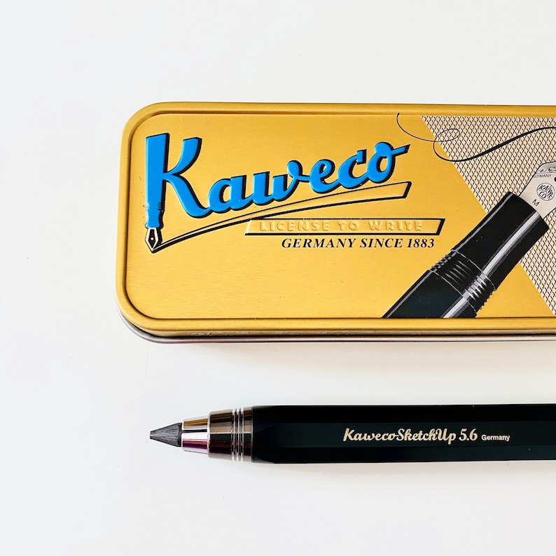 May include: A black mechanical pencil with a silver trim and the text "Kaweco SketchUp 5.6 Germany" on the side. The pencil is sitting on a white surface. The pencil is in a gold tin with the text "Kaweco License to Write Germany Since 1883" on it.