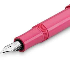 May include: Close-up of a pink fountain pen with a silver nib. The pen has a smooth, glossy finish and a classic design. The nib is detailed with intricate markings. The pen is set against a white background.