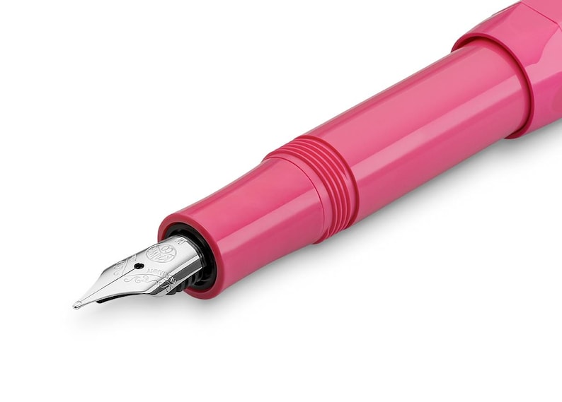 May include: Close-up of a pink fountain pen with a silver nib. The pen has a smooth, glossy finish and a classic design. The nib is detailed with intricate markings. The pen is set against a white background.