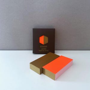 May include: Two stacks of colourful sticky notes in a brown box. The box has the text "De Paperie Doo Mimo" printed on it. The sticky notes are in orange and brown.