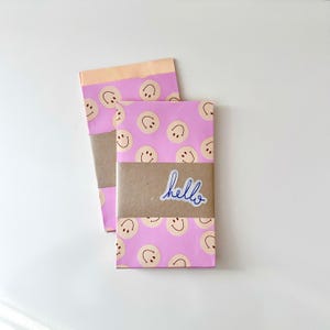 May include: Two wrapped packages with pink paper featuring smiley face patterns. One package is wrapped with brown paper and a sticker that says "hello". The other package is partially visible behind the first.