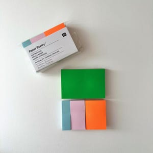 May include: A package of "Paper Poetry" sticky notes with text in English, German, and French. The package has a color block design with pink, orange, and blue. Loose sticky notes in green, blue, pink, and orange are also visible. The dimensions are 15x50mm and 25x50mm.