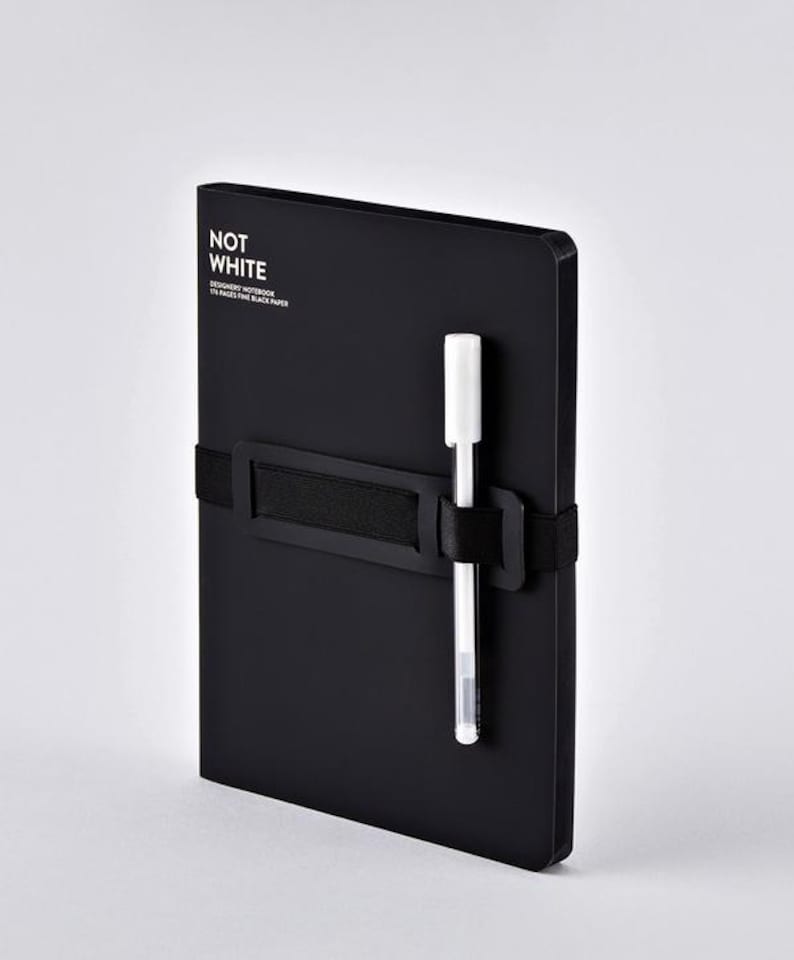 May include: A black notebook with the text "NOT WHITE" and a white pen secured by a black elastic band. The notebook is a designer's notebook with 176 pages of fine black paper.