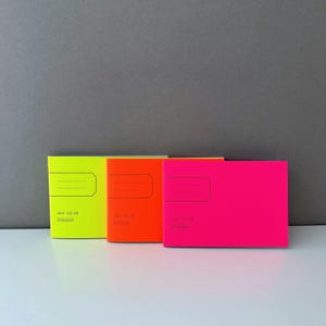 Könnte beinhalten: Drei kleine Notizbücher mit neonfarbenen Umschlägen. Die Notizbücher sind übereinander gestapelt. Das obere Notizbuch ist pink, das mittlere Notizbuch ist orange und das untere Notizbuch ist gelb. Die Notizbücher haben schwarzen Text auf der Vorderseite, der "48 P. 120 GR" und "Levenger" lautet.