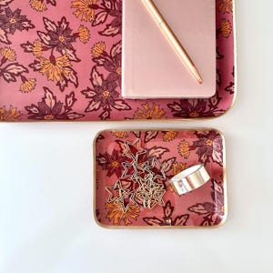 Könnte beinhalten: Zwei rechteckige Tabletts mit rosafarbenem Hintergrund und einem Blumenmuster in Orange-, Dunkelrot- und Violetttönen. Auf dem größeren Tablett liegen ein hellrosa Notizbuch und ein roségoldener Stift. Das kleinere Tablett enthält goldene Büroklammern.