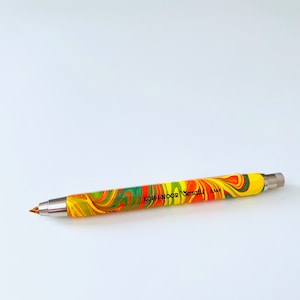 May include: A yellow, orange and green marbled mechanical pencil with a silver tip and the text "KOH-I-NOOR Versatil 5340" printed on the side.