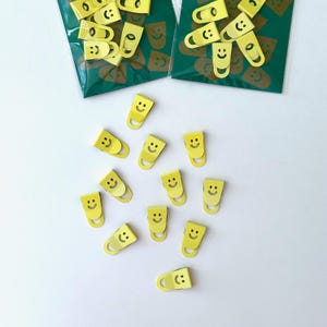 Può includere: Graffette gialle con un design a faccina sorridente sono sparse su una superficie bianca. Due confezioni delle stesse graffette sono visibili sullo sfondo, con uno sfondo verde e un motivo a faccina sorridente ripetuto.