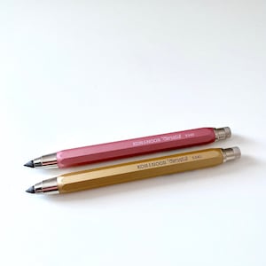 Könnte beinhalten: Zwei KOH-I-NOOR Versatile 5340 Druckbleistifte. Einer ist in Staubrosa, der andere in Goldgelb gehalten. Beide Stifte haben silberne Metallspitzen und Endkappen. Der Text "KOH-I-NOOR Versatile 5340" ist auf der Seite jedes Stifts aufgedruckt.