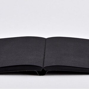 May include: An open, black notebook with a textured cover lies flat against a white background. The pages are also black, and the spine is visible. The notebook is centered and takes up most of the frame.