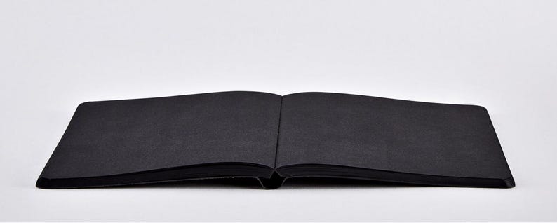 May include: An open, black notebook with a textured cover lies flat against a white background. The pages are also black, and the spine is visible. The notebook is centered and takes up most of the frame.