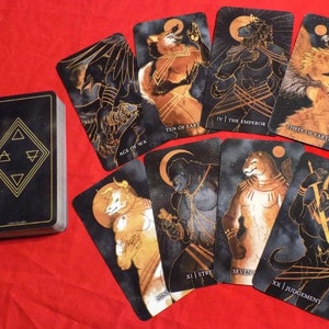 May include: A deck of tarot cards with black and gold designs. The cards feature illustrations of animals and mythical creatures. The cards are laid out on a red surface. The cards include the Ace of War, Ten of Earth, XI Strength, IV The Emperor, Three of Earth, Seven, and XX Judgement.