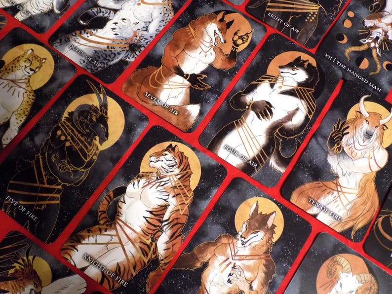 Puede incluir: Un juego de cartas de tarot con personajes de animales antropomorfos en varias poses. Las cartas son negras con detalles dorados y tienen un fondo estrellado. Las cartas incluyen "Ocho de Aire", "XII | El Ahorcado", "Reina de Aire", "Siete de Aire", "Cuatro de Aire", "Diez de Fuego", "Cinco de Fuego" y "Caballero de Fuego".
