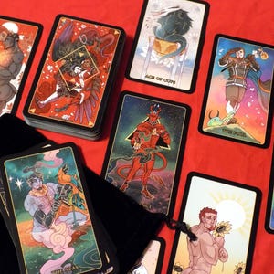 May include: A tarot deck with colorful illustrations on black cards. The cards feature a variety of characters and symbols, including a devil, a cup, a tower, and a star. The cards are arranged in a spread on a red surface.