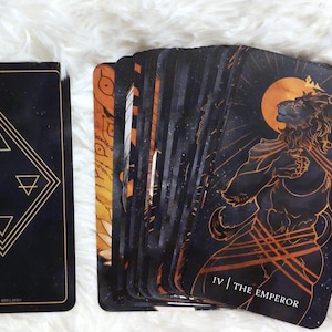 Puede incluir: Carta de tarot con fondo negro y detalles dorados. La carta representa una figura de le&oacute;n con una corona y un bast&oacute;n dorado. El texto "IV | THE EMPEROR" est&aacute; impreso en la parte inferior de la carta.