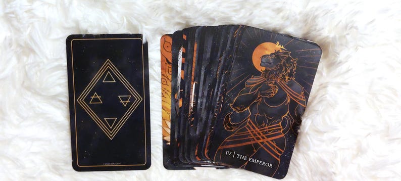 Puede incluir: Carta de tarot con fondo negro y detalles dorados. La carta representa una figura de le&oacute;n con una corona y un bast&oacute;n dorado. El texto "IV | THE EMPEROR" est&aacute; impreso en la parte inferior de la carta.