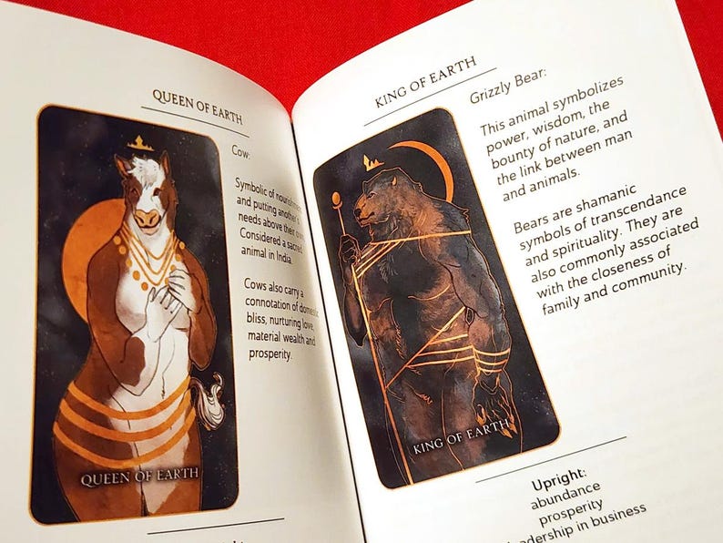 Puede incluir: Dos cartas de tarot, Reina de la Tierra y Rey de la Tierra, con ilustraciones de una vaca y un oso grizzly. La carta de la Reina de la Tierra tiene un texto que dice "Simb&oacute;lico de la nutrici&oacute;n y de poner las necesidades de los dem&aacute;s por encima de las propias. Considerado un animal sagrado en la India. Las vacas tambi&eacute;n llevan una connotaci&oacute;n de felicidad dom&eacute;stica, amor nutritivo, riqueza material y prosperidad." La carta del Rey de la Tierra tiene un texto que dice "Oso Grizzly: Este animal simboliza el poder, la sabidur&iacute;a, la abundancia de la naturaleza y el v&iacute;nculo entre el hombre y los animales. Los osos son s&iacute;mbolos cham&aacute;nicos de trascendencia y espiritualidad. Tambi&eacute;n se asocian com&uacute;nmente con la cercan&iacute;a de la familia y la comunidad." El texto de la carta est&aacute; en marr&oacute;n.