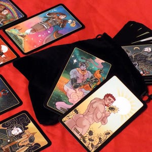 May include: A set of tarot cards with colorful illustrations and gold accents. The cards are spread out on a red surface with a black velvet bag and a stack of cards in the background.
