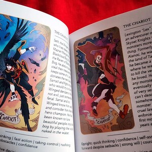 Pu&ograve; includere: Libro aperto con due illustrazioni di un mazzo di tarocchi fantasy. Uno mostra una figura alata in nero e arancione, etichettata "Il Carro". L'altro raffigura una figura dai capelli rossi con una spada, anch'essa "Il Carro".