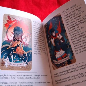Pu&ograve; includere: Libro aperto con due illustrazioni di carte dei tarocchi. Una carta presenta una figura dalla pelle blu con le corna che tiene una spada, etichettata come "ACE OF SWORDS". L'altra carta mostra una figura con una spada e dettagli rossi. Il testo descrive i significati delle carte.