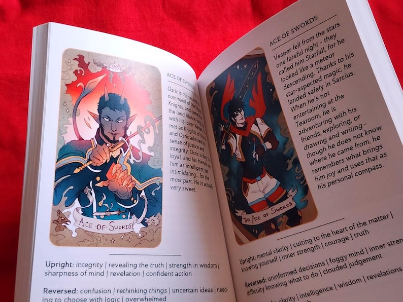 Pu&ograve; includere: Libro aperto con due illustrazioni di carte dei tarocchi. Una carta presenta una figura dalla pelle blu con le corna che tiene una spada, etichettata come "ACE OF SWORDS". L'altra carta mostra una figura con una spada e dettagli rossi. Il testo descrive i significati delle carte.
