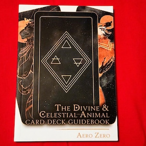 Puede incluir: Una gu&iacute;a blanca con una ilustraci&oacute;n de baraja de cartas negra y dorada en la portada. El t&iacute;tulo es "The Divine & Celestial Animal Card Deck Guidebook" con el nombre del autor "Aero Zero" en la parte inferior.