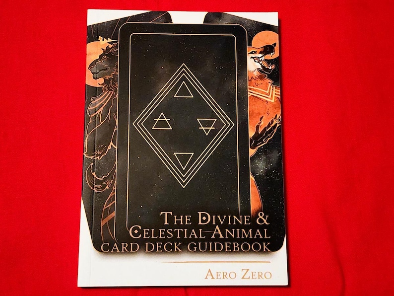 Puede incluir: Una gu&iacute;a blanca con una ilustraci&oacute;n de baraja de cartas negra y dorada en la portada. El t&iacute;tulo es "The Divine & Celestial Animal Card Deck Guidebook" con el nombre del autor "Aero Zero" en la parte inferior.